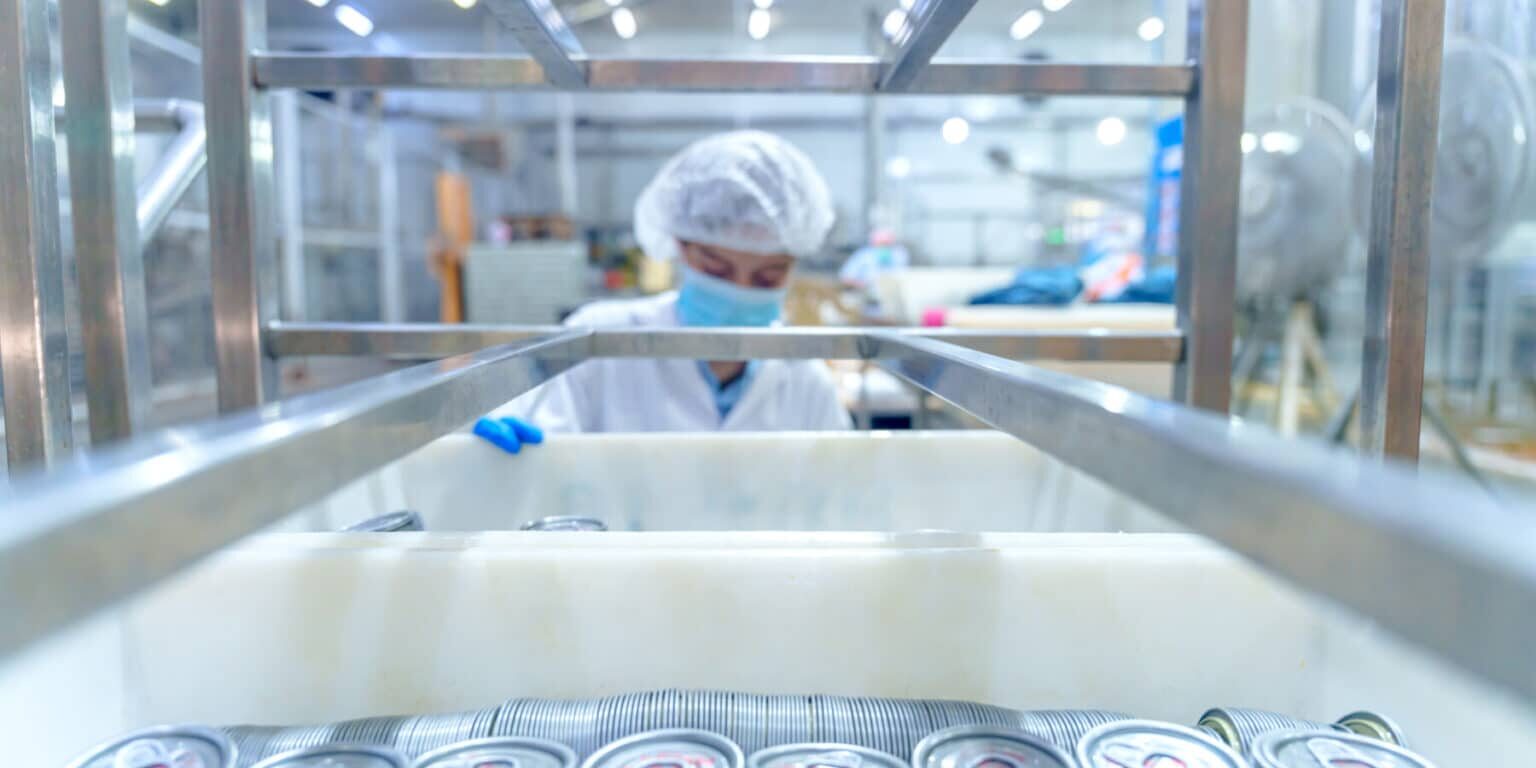 A worker in protective clothing and mask stands in a factory behind a rack of stacked metal cans, with bright lights overhead, ensuring pet food supplier qualification standards are strictly maintained.