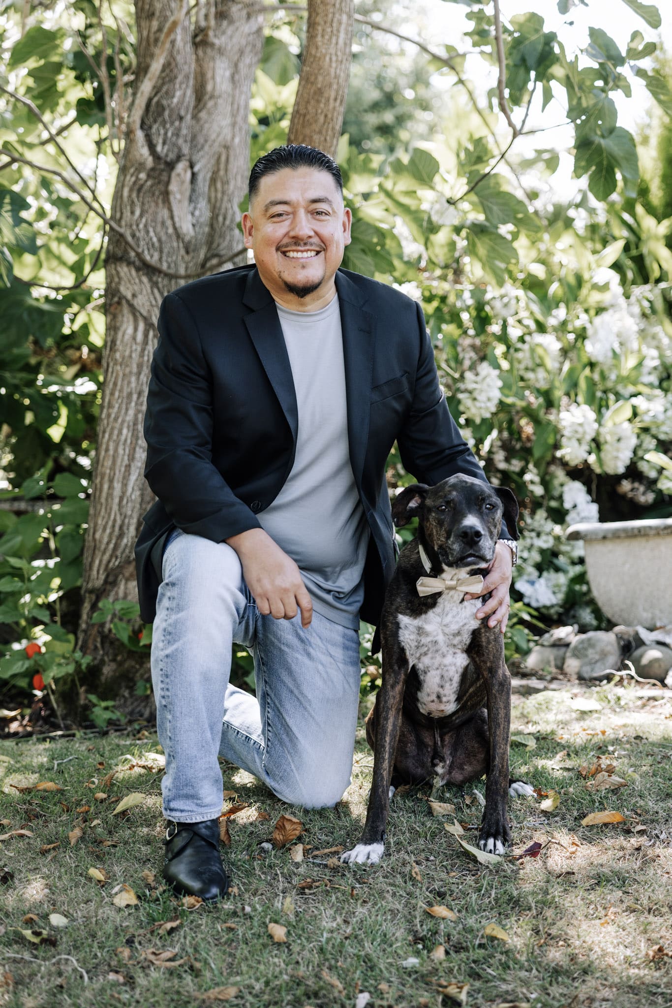 Man kneeling on grass next to a sitting dog, outdoors with trees and plants in the background. The man is smiling and wearing a blazer and jeans, enjoying some downtime after overseeing pet food operations.