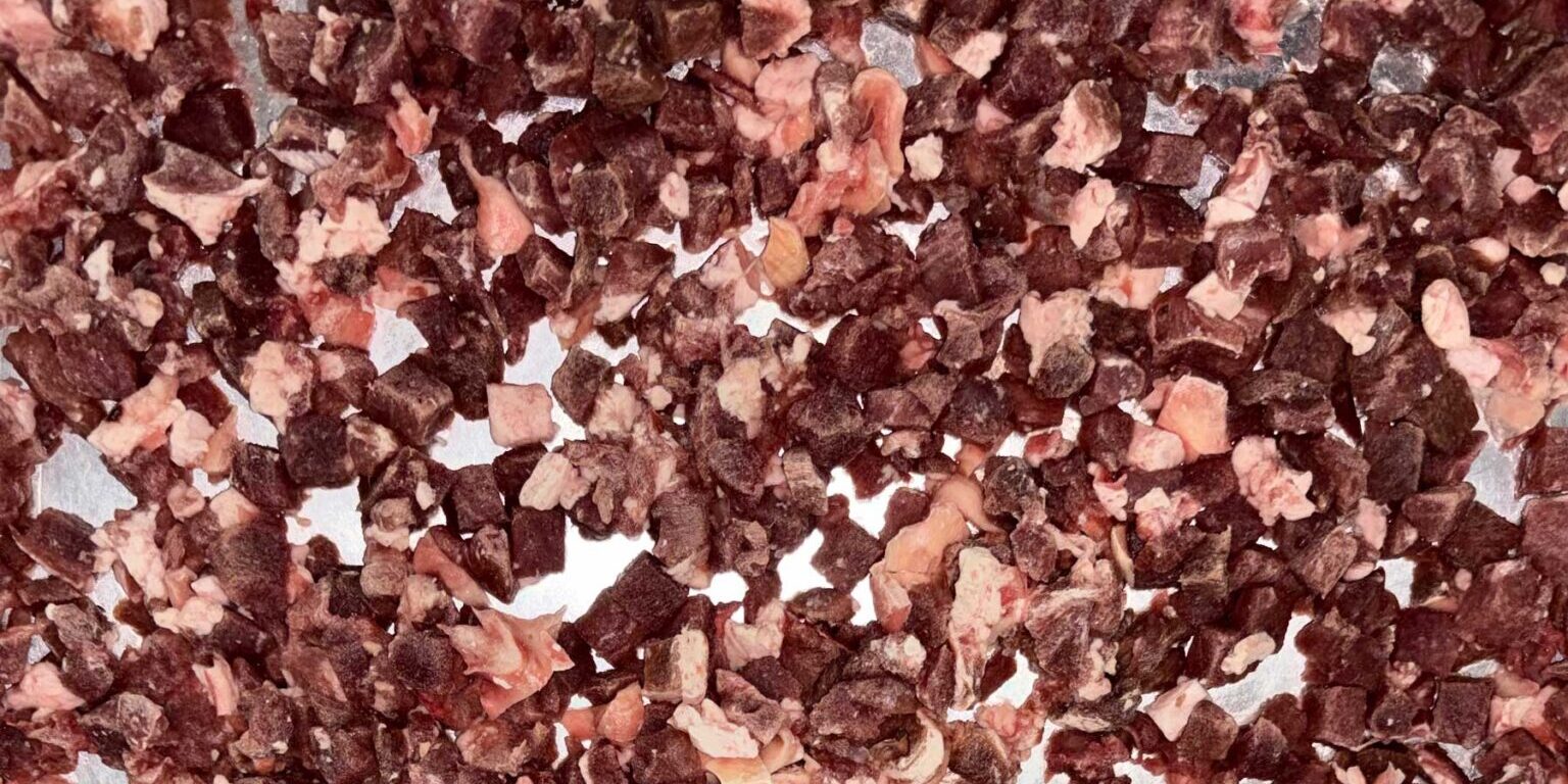 Small, irregular pieces of dried red meat—sourced with care through responsible pet food ingredient sourcing—are scattered closely together on a flat surface.