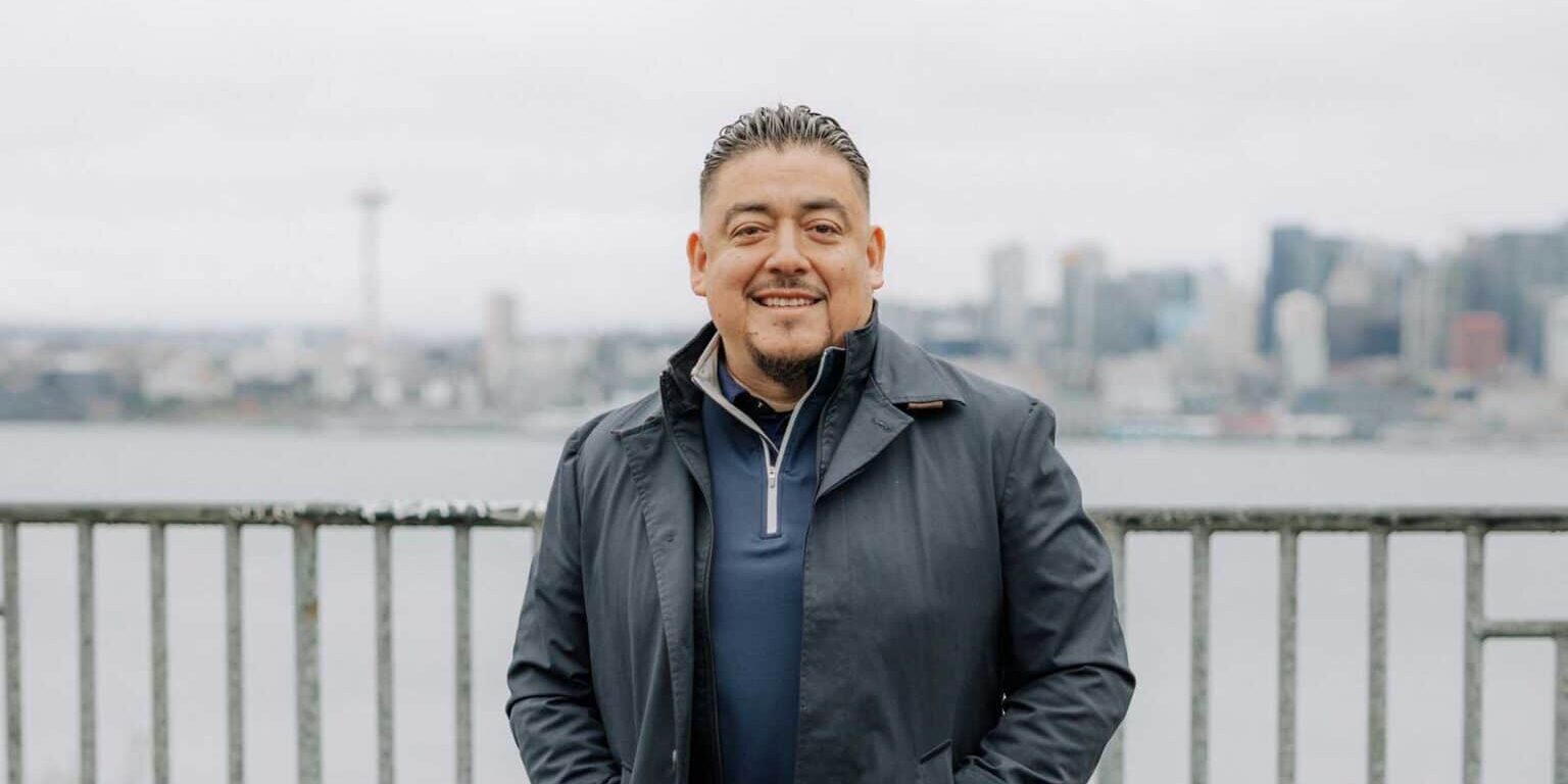 Man in a dark coat standing in front of a metal railing with a city skyline and overcast sky in the background, exuding the calm confidence of a seasoned pet food operations leader.